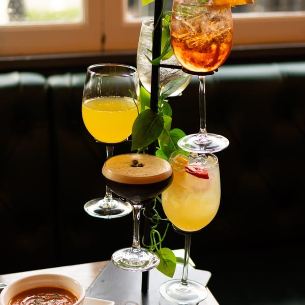 A drink stand with five cocktails, a bowl of soup, and bao buns on a table.