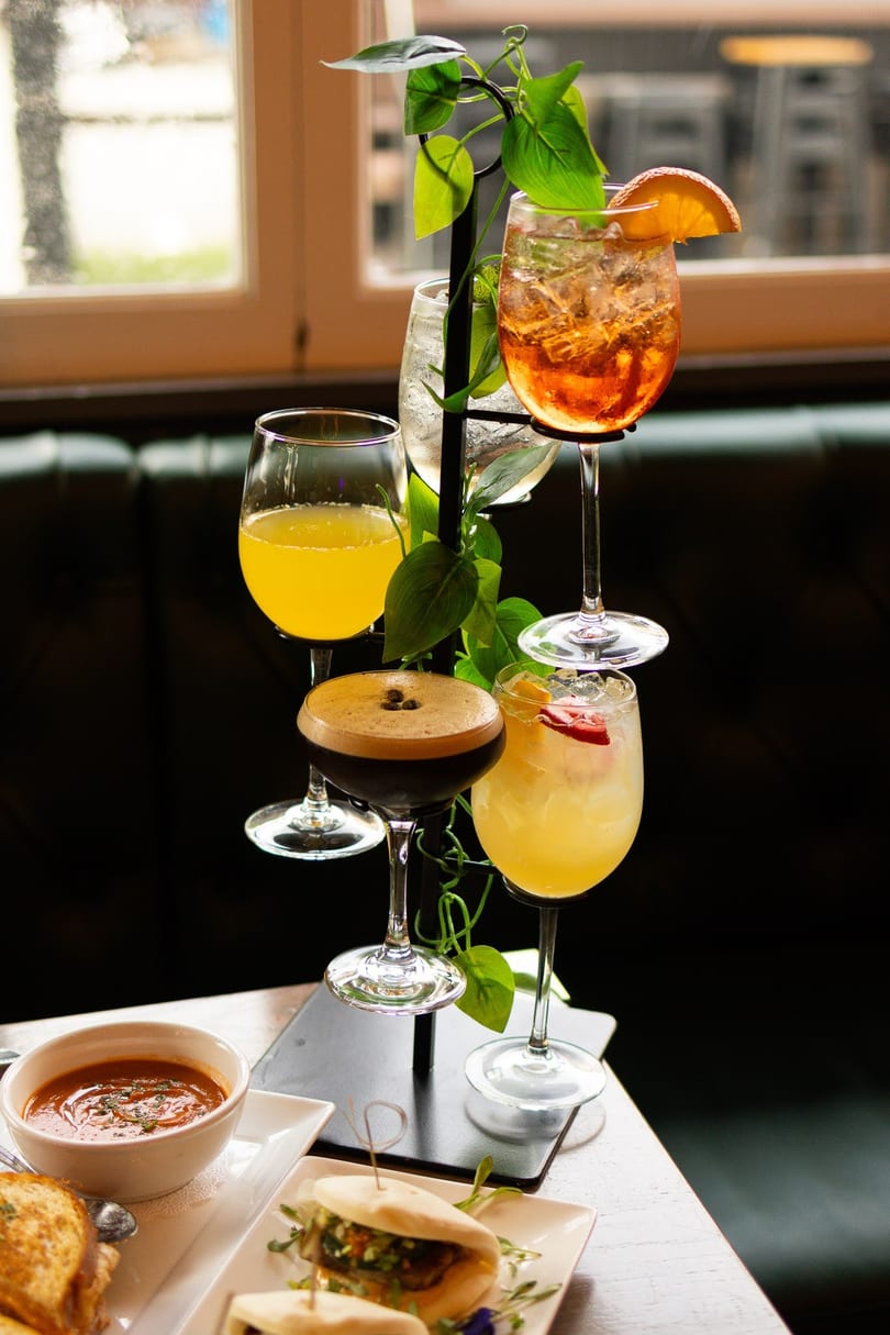 A drink stand with five cocktails, a bowl of soup, and bao buns on a table.
