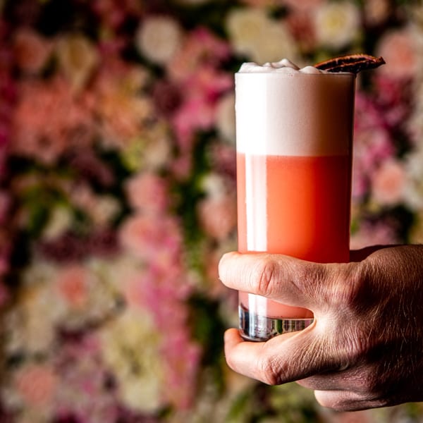 Hand holding a vibrant pink cocktail with foamy top and garnish against a blurred floral wall.