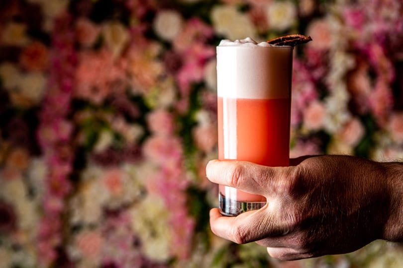 Hand holding a vibrant pink cocktail with foamy top and garnish against a blurred floral wall.