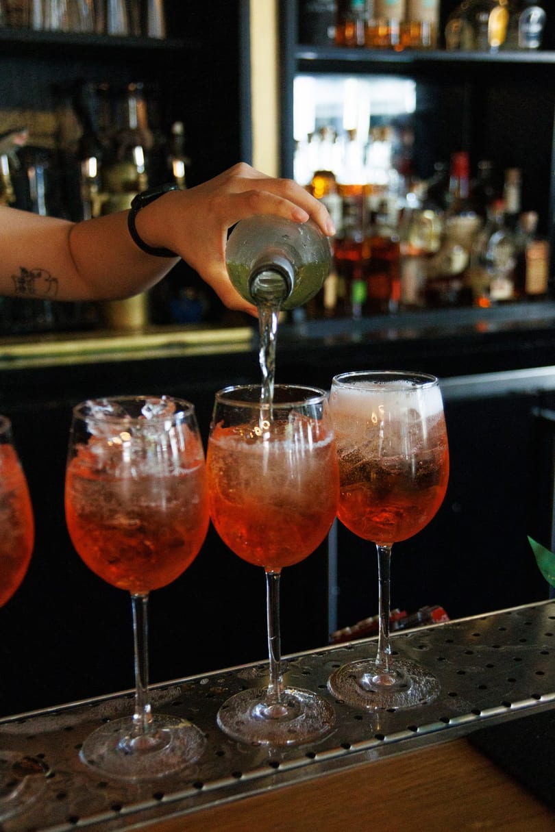 A bartender pours a drink from a bottle into a glass of orange cocktails on a bar.