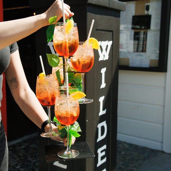 A server carries a tiered tray of five orange cocktails with lemon slices and straws.