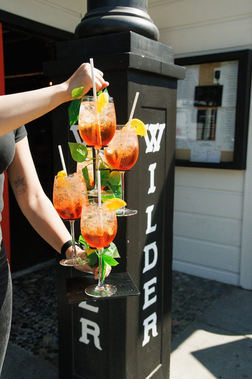 A server carries a tiered tray of five orange cocktails with lemon slices and straws.