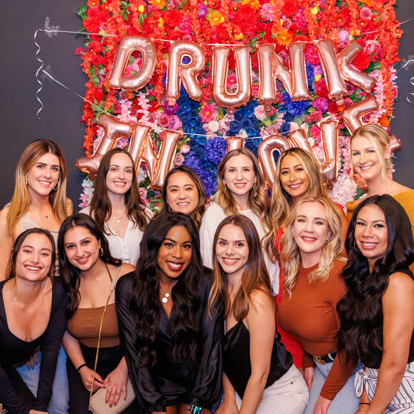 Group of smiling women posing in front of a colorful floral wall with "DRUNK IN LOVE" balloons.