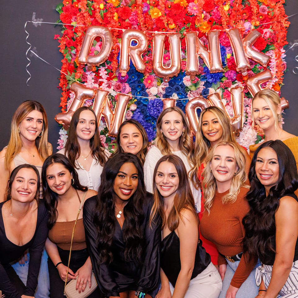 Group of smiling women posing in front of a colorful floral wall with "DRUNK IN LOVE" balloons.
