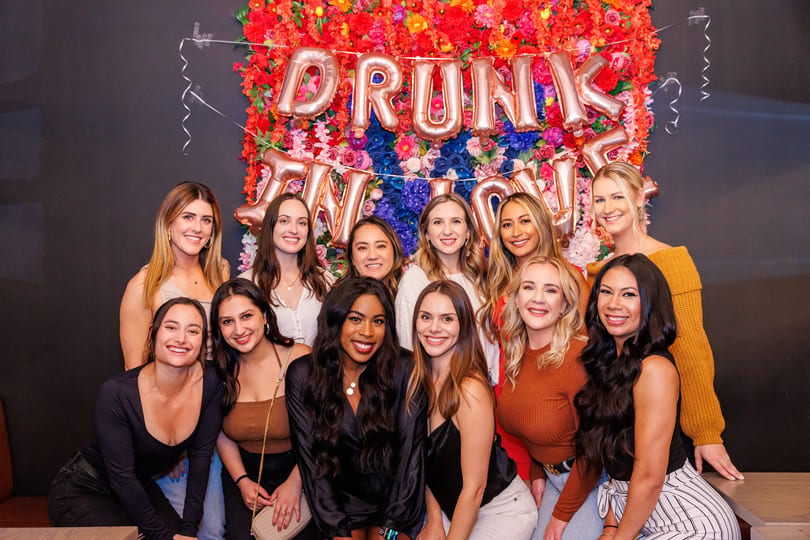 Group of smiling women posing in front of a colorful floral wall with "DRUNK IN LOVE" balloons.