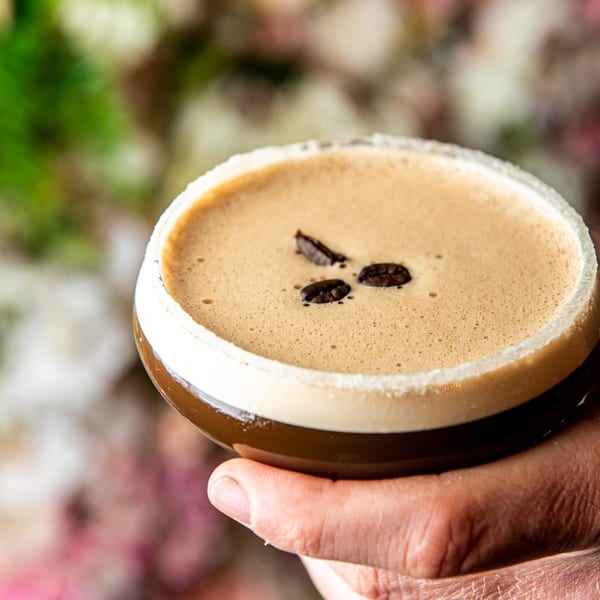 A hand holds an espresso martini garnished with coffee beans against a floral background.