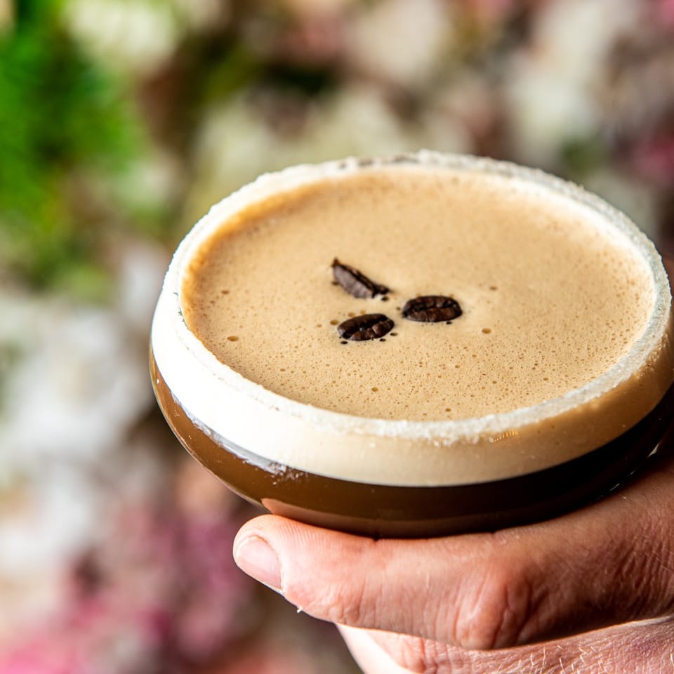 A hand holds an espresso martini garnished with coffee beans against a floral background.
