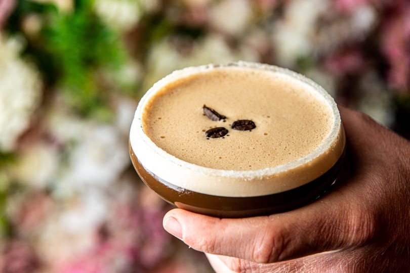 A hand holds an espresso martini garnished with coffee beans against a floral background.