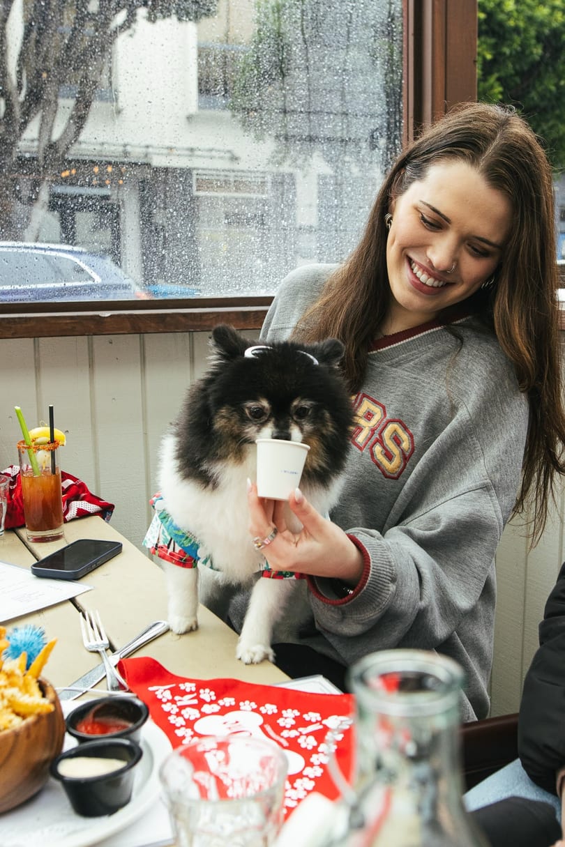 A smiling woman holds a small white cup for a fluffy black and white dog at a restaurant.