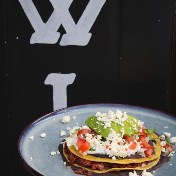 A plate of stacked tostadas with black beans, meat, avocado, tomatoes, and crumbled cheese.
