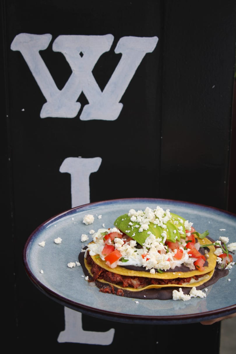 A plate of stacked tostadas with black beans, meat, avocado, tomatoes, and crumbled cheese.