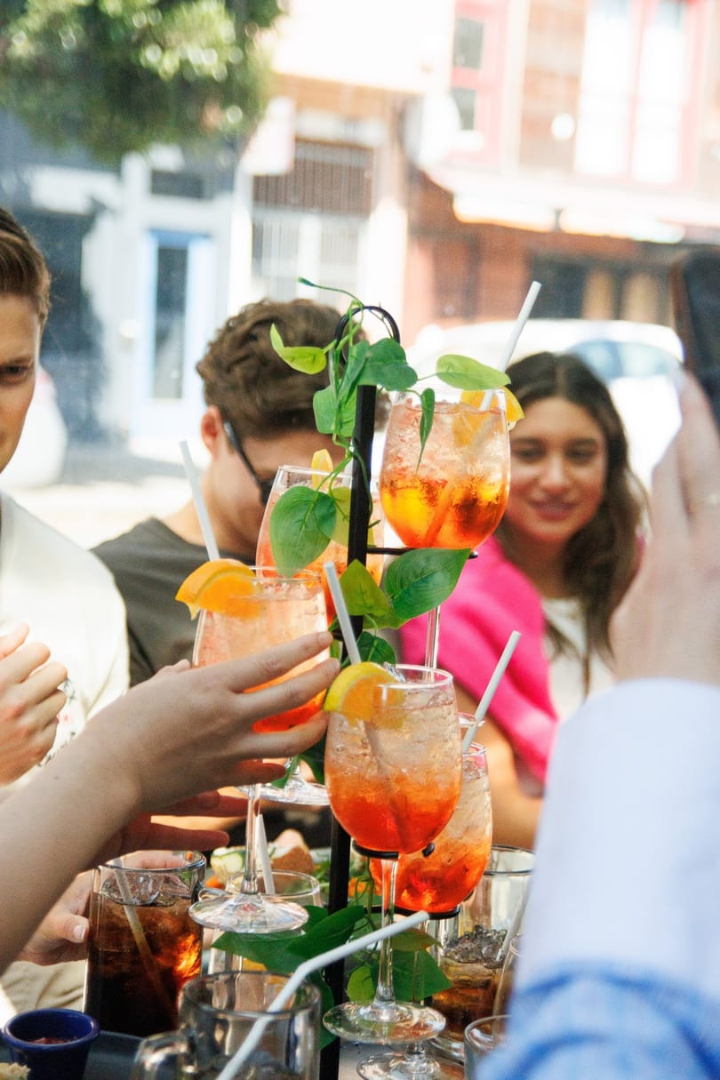 Friends enjoying orange cocktails with straws and orange slices at an outdoor restaurant.