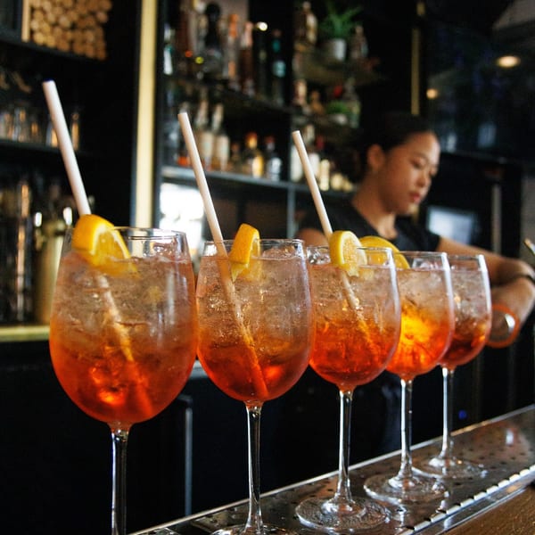 Five Aperol Spritz cocktails with orange slices and straws lined up on a bar, with a bartender in the background.