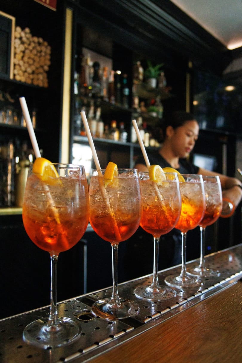 Five Aperol Spritz cocktails with orange slices and straws lined up on a bar, with a bartender in the background.