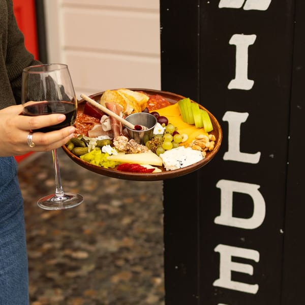 A person holds a charcuterie board with meats, cheeses, fruit, and nuts, and a glass of red wine.
