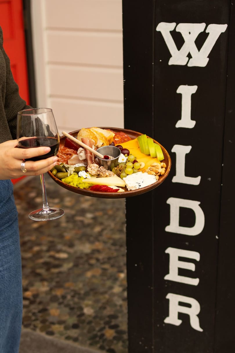 A person holds a charcuterie board with meats, cheeses, fruit, and nuts, and a glass of red wine.