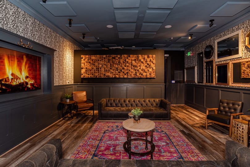 Dark, cozy lounge with leather seating, a fireplace screen, colorful rug, and wooden wall art.